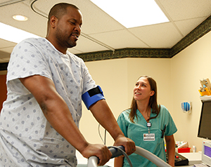 Man with wires attached to chest walking on treadmill. Health care provider is taking man's blood pressure.